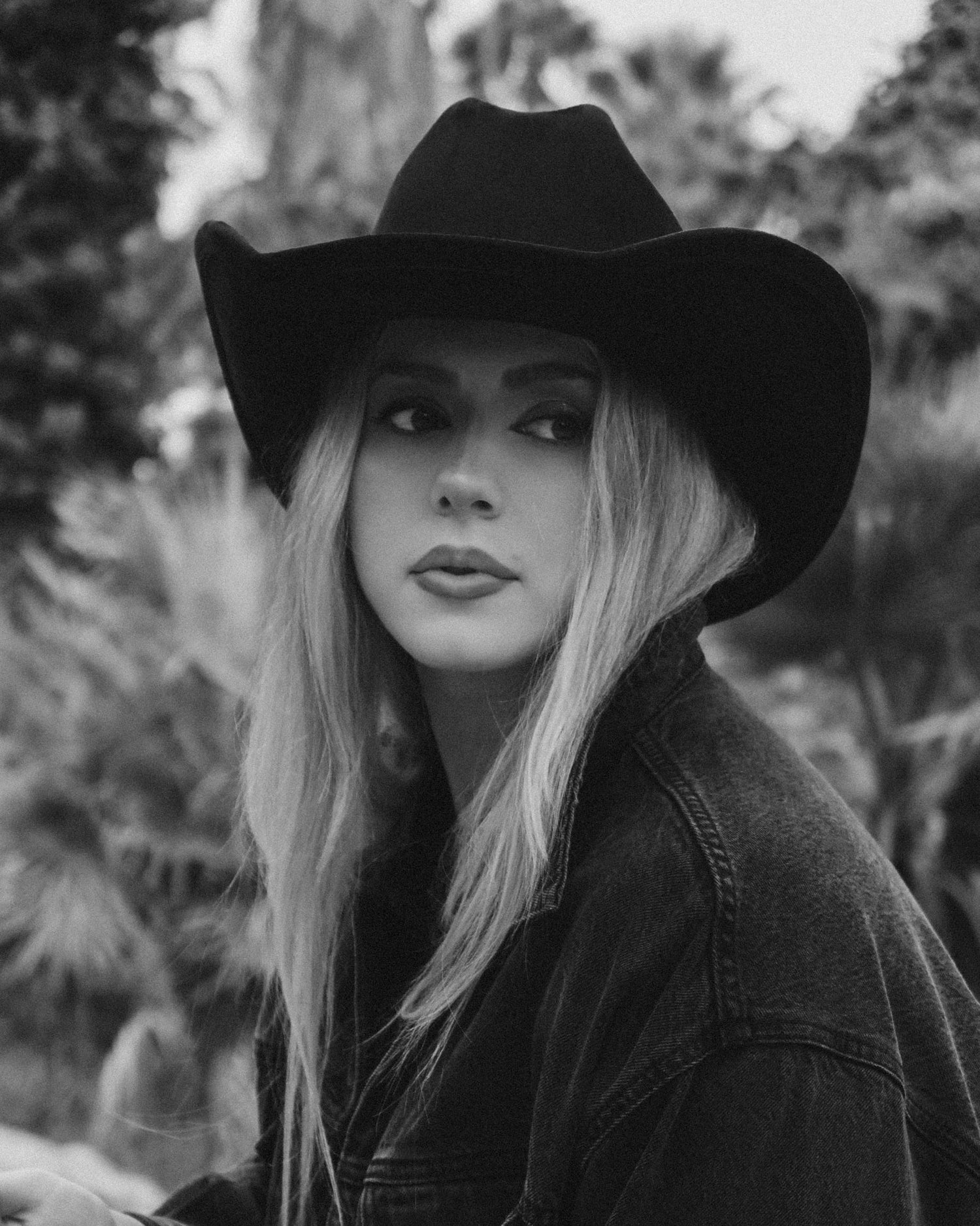 Black and white photo of a person wearing a wide-brimmed hat and dark coat outdoors.